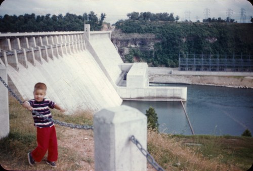 A young boy in a striped shirt and red pants plays near a large dam and river, with power lines in the background.