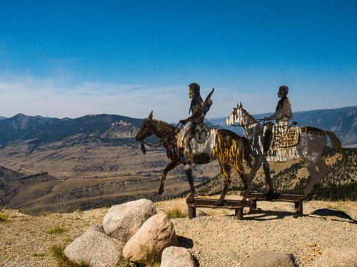 Metal sculptures of two riders on horseback overlook a mountainous landscape under a clear blue sky.