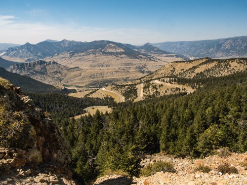 A panoramic view of rugged mountains and a winding dirt road, surrounded by dense green forests under a clear blue sky.