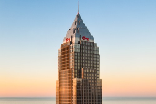 A tall skyscraper with a peaked roof and a logo at the top, set against a clear sky and ocean backdrop.