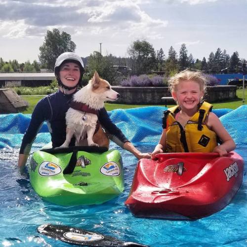 A child and an adult in kayaks, smiling, with a corgi sitting on the adult's lap, against a sunny outdoor backdrop.