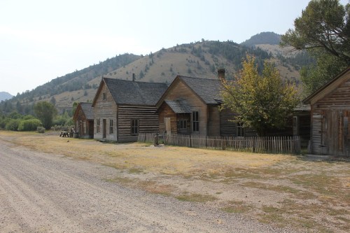 Abandoned wooden buildings along a gravel road, with mountains in the background and sparse vegetation.
