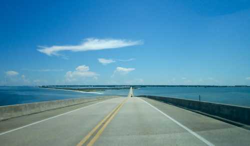 A long road stretches over water, leading towards a distant shoreline under a clear blue sky with a few clouds.