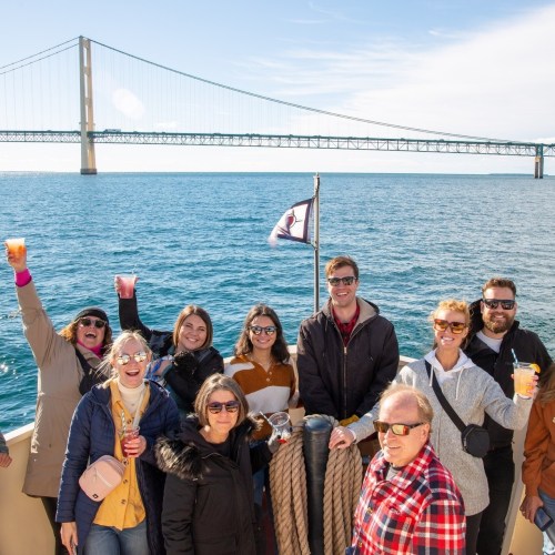 A group of people smiling and posing on a boat with a bridge in the background on a sunny day.