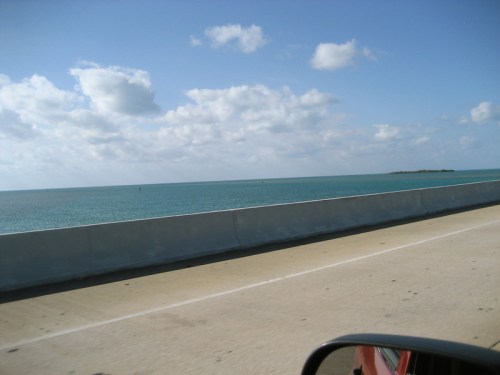 A scenic view of the ocean from a highway, with blue water and fluffy clouds under a bright sky.
