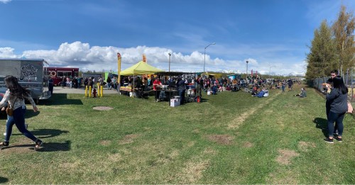 A bustling outdoor market with food trucks, tents, and crowds enjoying a sunny day on green grass.