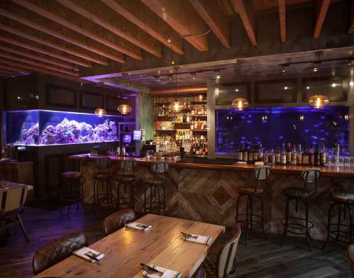 A stylish bar area with a large aquarium, wooden tables, and warm lighting, creating a cozy atmosphere.