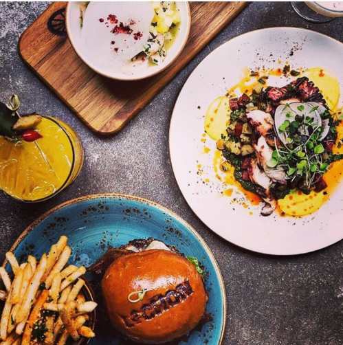 A vibrant spread featuring a burger, fries, seafood dish, and a cocktail on a textured surface.