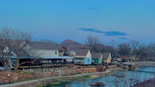 A serene lakeside view featuring quaint houses, trees, and a distant mountain under a blue sky at dusk.