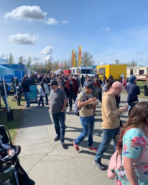 A busy outdoor food truck event with crowds of people enjoying food and a clear blue sky.