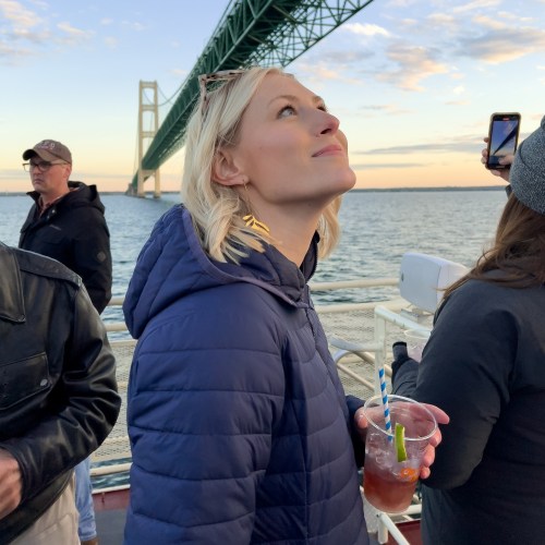 A woman in a blue hoodie looks up, holding a drink, with a bridge and water in the background.