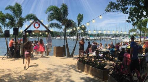 A lively beach scene with people enjoying a waterfront event, surrounded by palm trees and boats in the background.