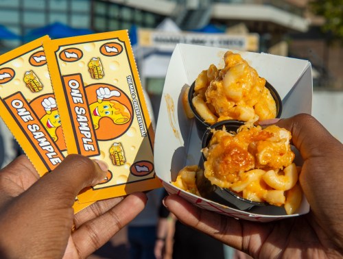 Two hands hold a container of cheesy macaroni and cheese with sample tickets in the background.