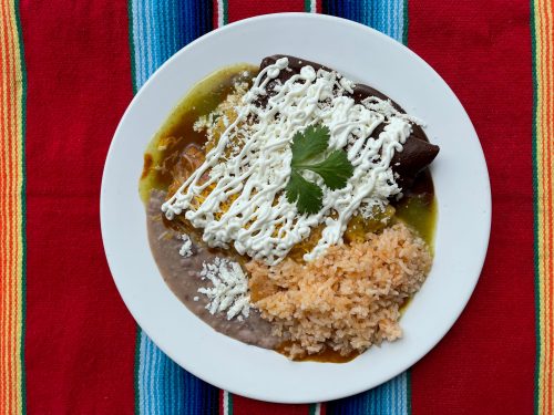 A plate of enchiladas topped with sour cream and cilantro, served with rice and refried beans on a colorful tablecloth.