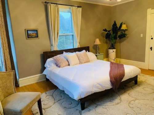 Cozy bedroom with a double bed, decorative pillows, a chair, and a plant, featuring soft lighting and warm colors.