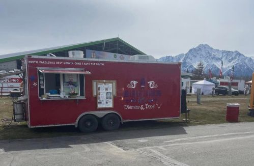 A red food truck with a menu, set against a backdrop of mountains and a cloudy sky.