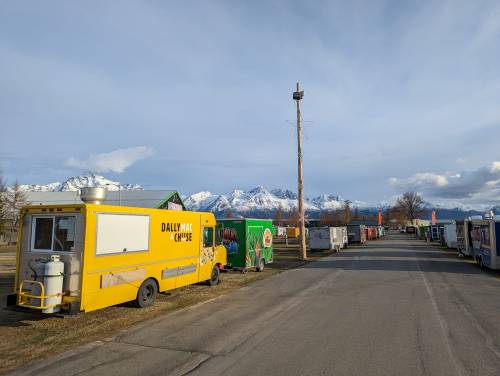 A row of colorful food trucks lined up along a road with snow-capped mountains in the background under a clear sky.