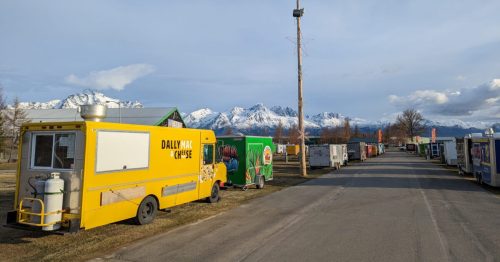 A row of colorful food trucks lined up on a street with snow-capped mountains in the background.