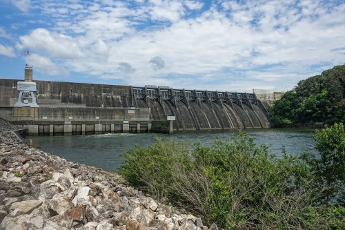 A large dam with water flowing in front, surrounded by greenery and a cloudy sky.