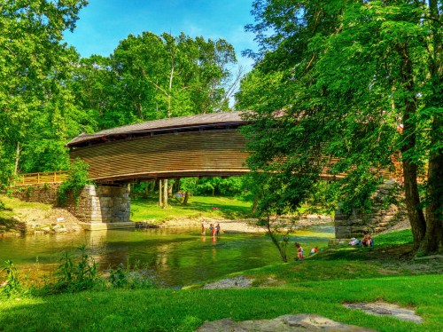 A wooden covered bridge spans a serene river, surrounded by lush green trees and people enjoying the water nearby.