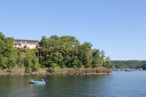 A serene lake scene with two fishermen in a boat, surrounded by lush greenery and a house on a hillside.