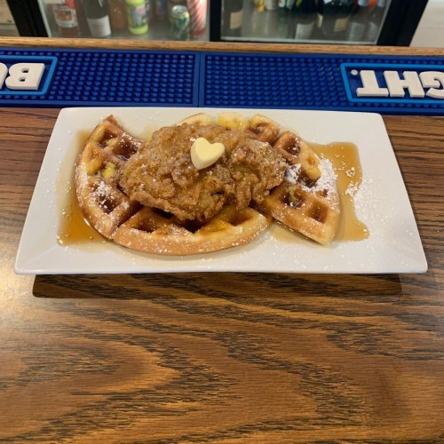 A plate of chicken and waffles drizzled with syrup, topped with a small heart-shaped butter pat.