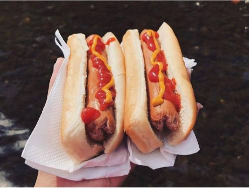 A hand holding two hot dogs in buns, topped with ketchup and mustard, against a blurred outdoor background.