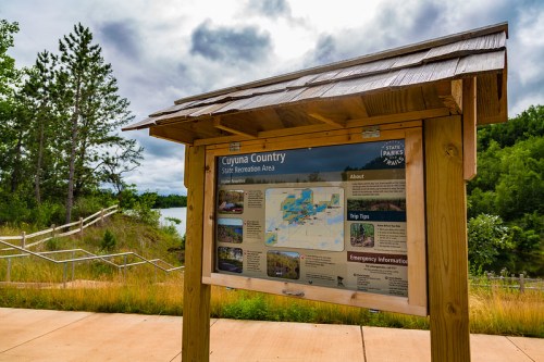 Information sign for Cuyuna Country State Recreation Area, featuring maps and tips, surrounded by greenery and a lake.