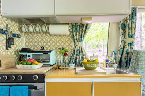 Bright kitchen with patterned wallpaper, a stove, sink, and colorful fruits in bowls on a yellow countertop.