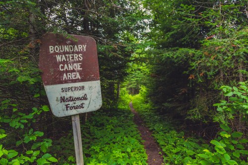 A weathered sign marks the Boundary Waters Canoe Area, surrounded by lush greenery and a narrow forest path.