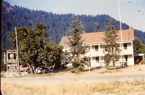 A vintage roadside diner with a sign, surrounded by trees and mountains in the background. Classic cars parked nearby.