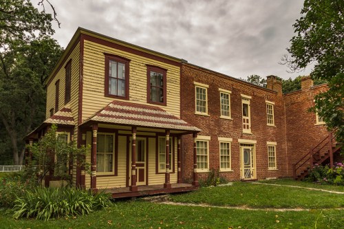 A historic two-story building with yellow and red brick exterior, surrounded by greenery under a cloudy sky.