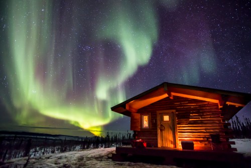 A cozy wooden cabin under a vibrant aurora borealis and starry night sky. Snow covers the ground.