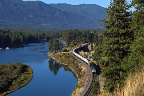 A train winds along a riverbank, surrounded by lush trees and mountains under a clear blue sky.