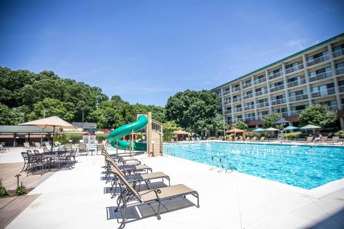 A sunny pool area with a water slide, lounge chairs, and a hotel building in the background surrounded by trees.