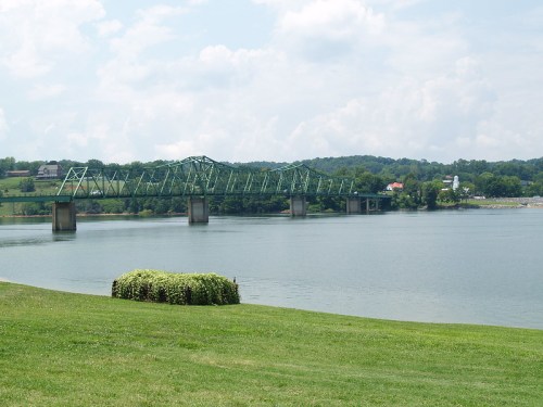 A green bridge spans a calm river, with grassy banks and a cloudy sky in the background.