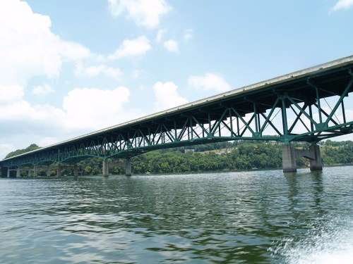 A green steel bridge spans over a calm river, with trees lining the banks under a blue sky with fluffy clouds.