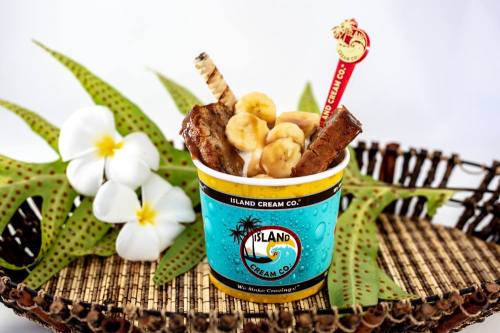 A colorful ice cream cup topped with banana slices and chocolate sticks, surrounded by tropical decor and flowers.