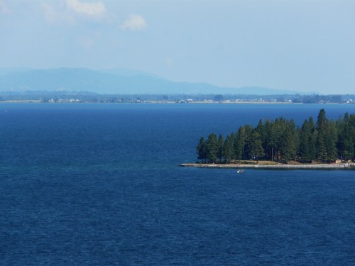 A serene blue lake with a small island of pine trees in the foreground and distant mountains on the horizon.