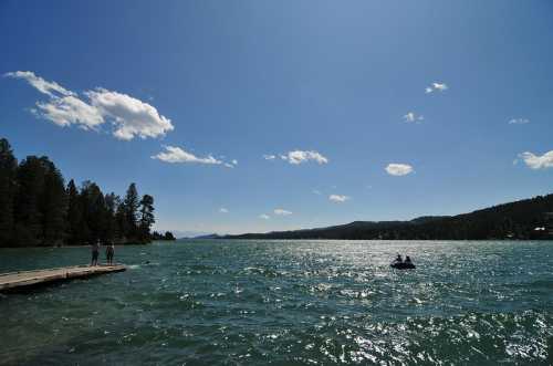 A serene lake scene with a wooden dock, people on the shore, and a small boat on the water under a clear blue sky.