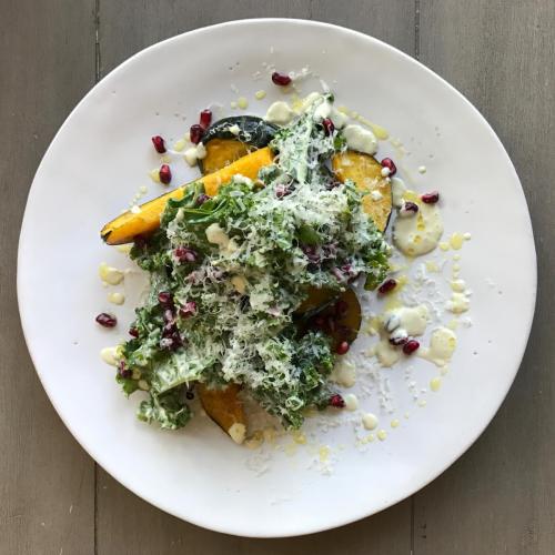 A vibrant salad with greens, roasted squash, pomegranate seeds, and a drizzle of creamy dressing, served on a white plate.