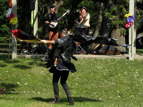 A performer in a black outfit dances with flowing fabric, while musicians play in the background at an outdoor event.