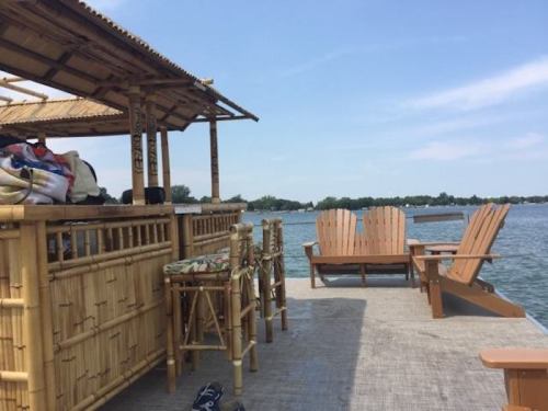 A bamboo deck overlooking a calm lake, featuring wooden chairs and a bar area under a thatched roof.
