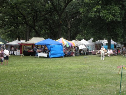 A vibrant outdoor market with colorful tents and people browsing in a grassy park setting.