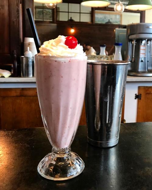A tall glass of strawberry milkshake topped with whipped cream and a cherry, next to a metal shaker.