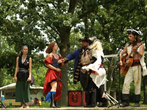 A lively outdoor scene with performers in pirate costumes, singing and acting on stage, surrounded by trees.