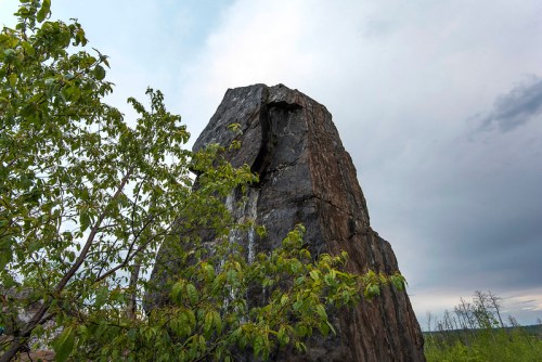 A tall, rugged rock formation surrounded by green foliage under a cloudy sky.