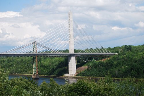 A modern cable-stayed bridge spans a river, surrounded by lush greenery and a cloudy sky.
