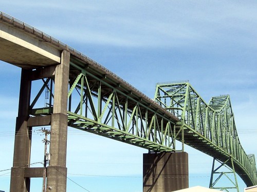 A green steel bridge spans across a blue sky, showcasing its intricate truss design and supporting pillars.