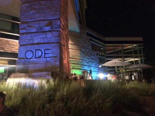 A stone building at night with colorful lighting, featuring the word "ODE" and outdoor seating under umbrellas.